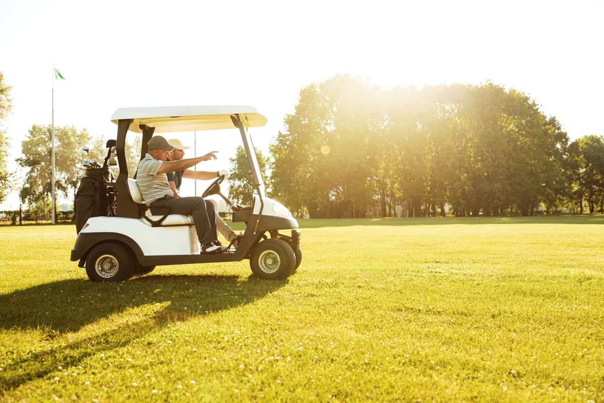 Two golfers riding in a golf cart, one pointing down the sunlit fairway